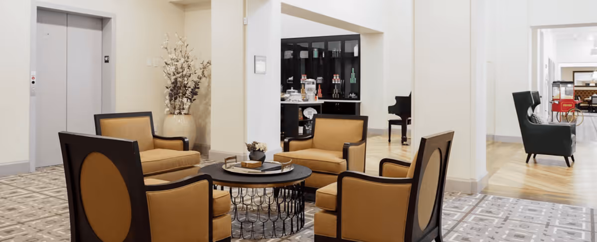 A seating area in a senior living facility with four mustard-colored armchairs arranged around a round black coffee table. In the background, there is an elevator door, a large vase with decorative branches, a black cabinet with glass doors displaying items, and a black piano. The floor has patterned tiles and wood flooring in different sections.