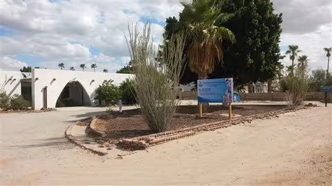 Desert landscaping frames a white, arched building entrance with a blue sign.