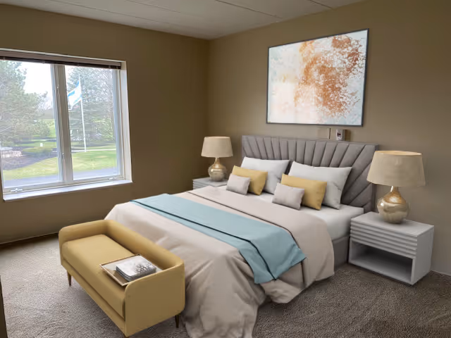 A neatly made bed with beige and light blue bedding in a bedroom. The bed has a padded headboard and is flanked by two bedside tables, each with a lamp. A small bench with a book on it is placed at the foot of the bed. There is a large window to the left showing a green outdoor area with trees and a flag. A large abstract painting hangs on the wall above the bed.