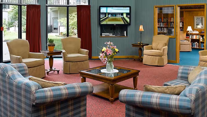 A cozy living room area with plaid upholstered sofas and beige armchairs arranged around a wooden coffee table with a vase of flowers. The room has large windows with red curtains, a wall-mounted TV displaying a bowling game, and an adjacent room with bookshelves and additional seating.