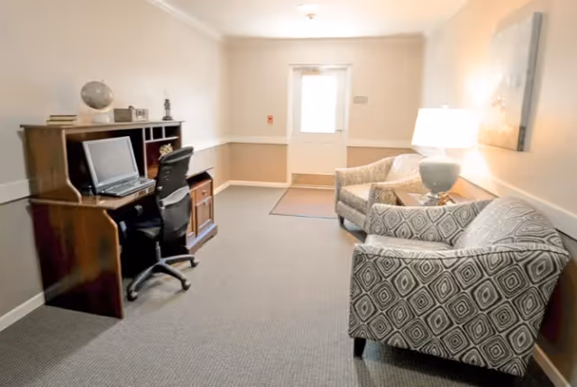 Small communal lounge with a wooden desk and computer on the left, two patterned armchairs and a lamp on the right, and a door at the far end.