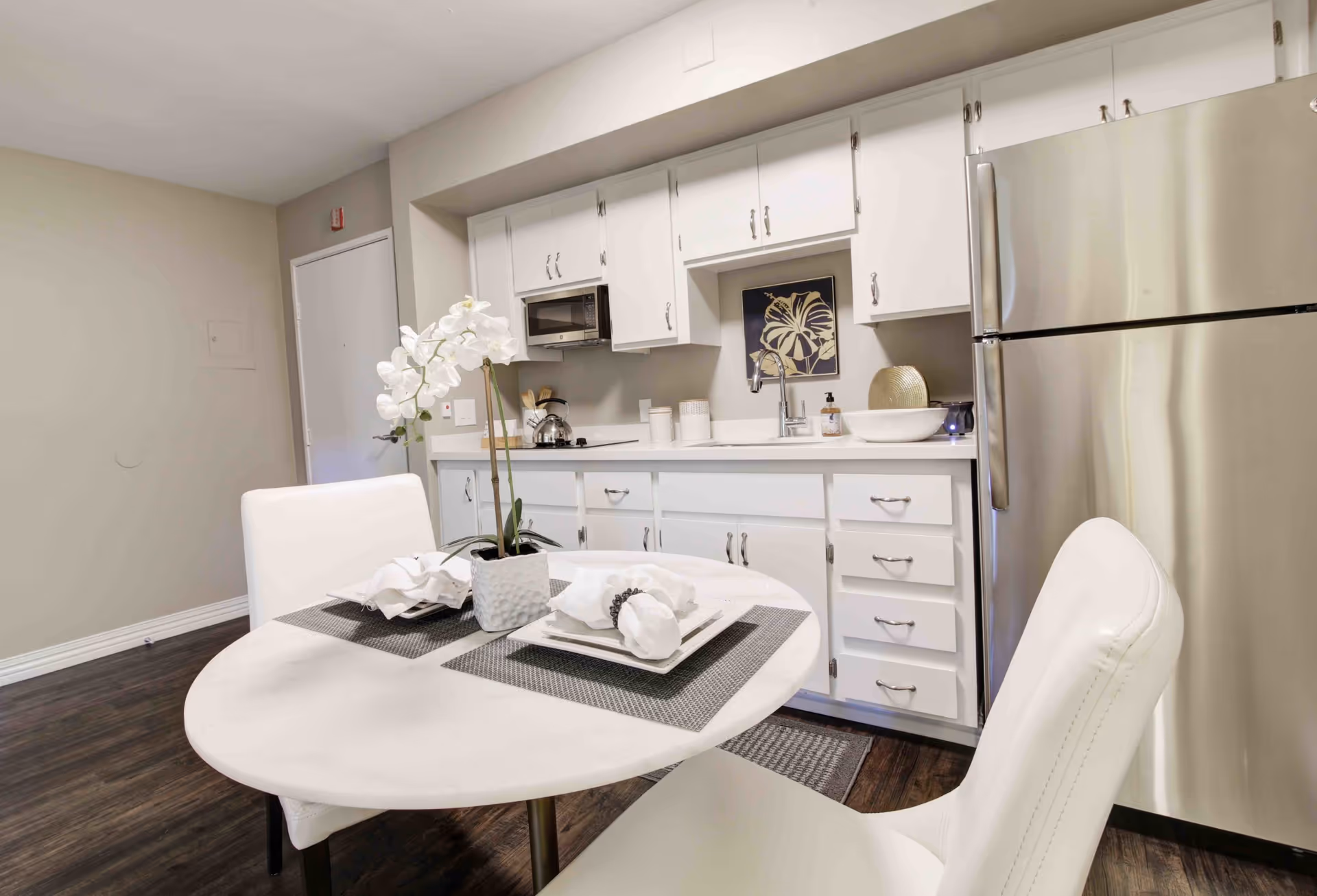 A modern kitchen area with white cabinets, a stainless steel refrigerator, a microwave, and a sink. In the foreground, there is a round white table set with two place settings and a white orchid centerpiece. The floor is dark wood, and the walls are painted light gray.
