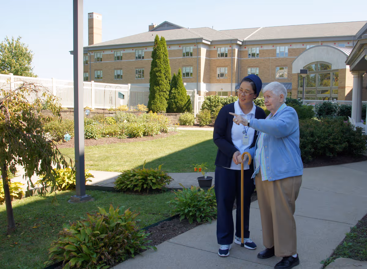 An elderly woman with a cane and a caregiver standing and pointing together in a landscaped courtyard outside a senior living building.