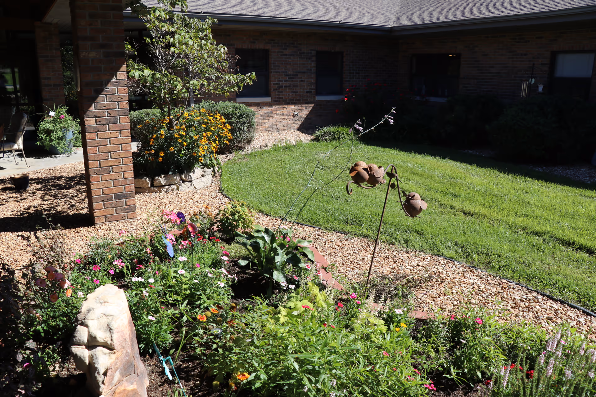 A garden area with various colorful flowers and plants, a small tree, and decorative metal bird ornaments. The garden is bordered by a gravel path and is adjacent to a brick building with windows. There is a covered patio area with chairs visible on the left side.