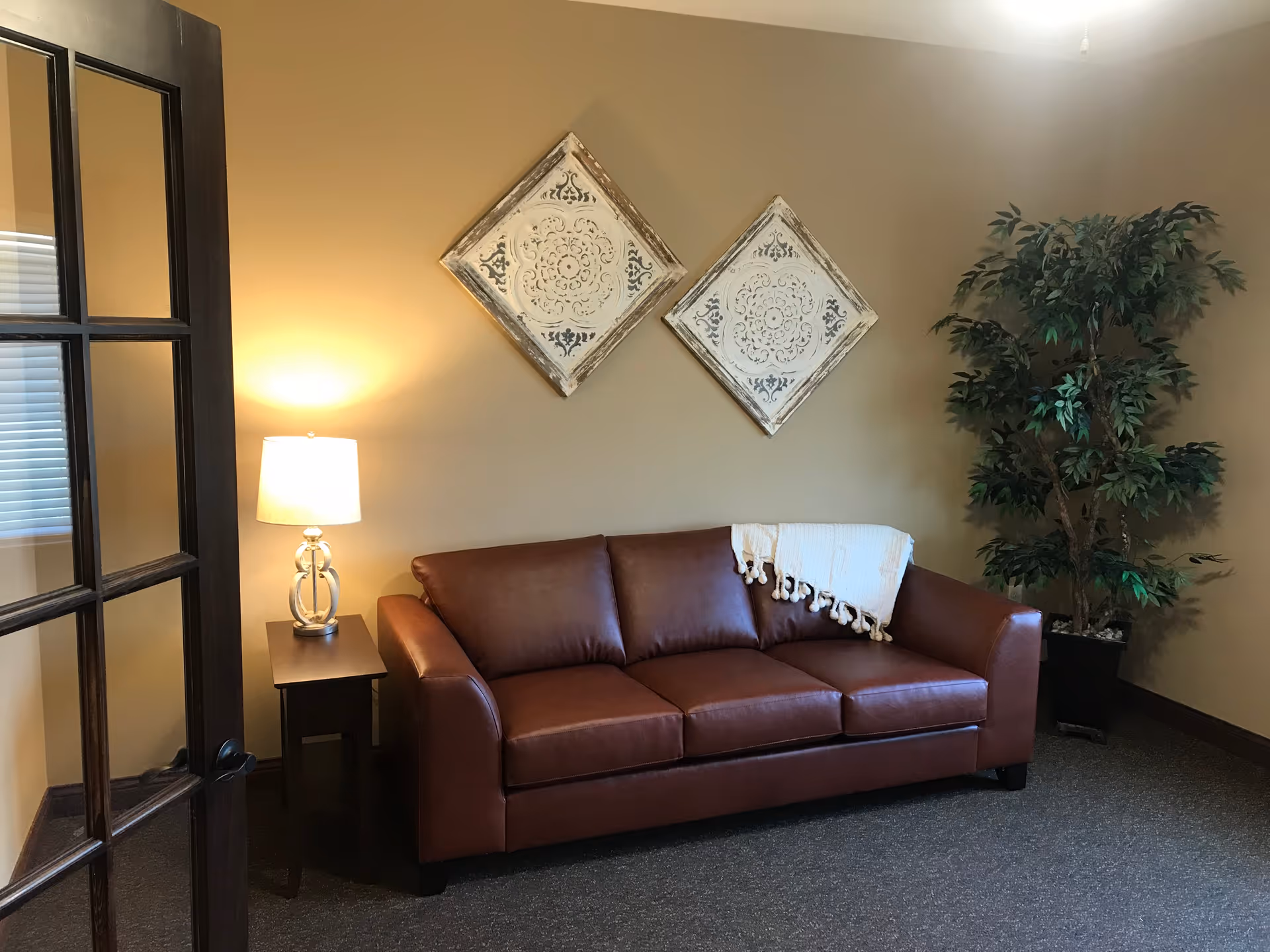 A cozy living room area with a brown leather sofa, a white throw blanket draped over one arm, a small side table with a lamp, two decorative square wall hangings, and a large potted plant in the corner. The room has beige walls and a glass-paneled door partially open.