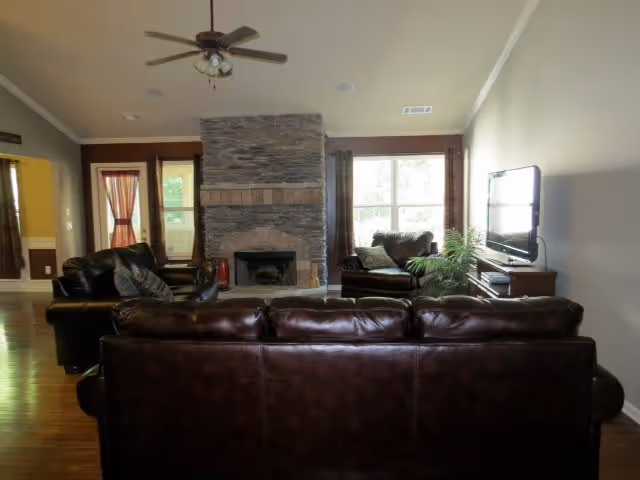 A living room with dark brown leather sofas arranged around a stone fireplace. There is a ceiling fan above, large windows with curtains on either side of the fireplace, a flat-screen TV mounted on the right wall, and a wooden floor.