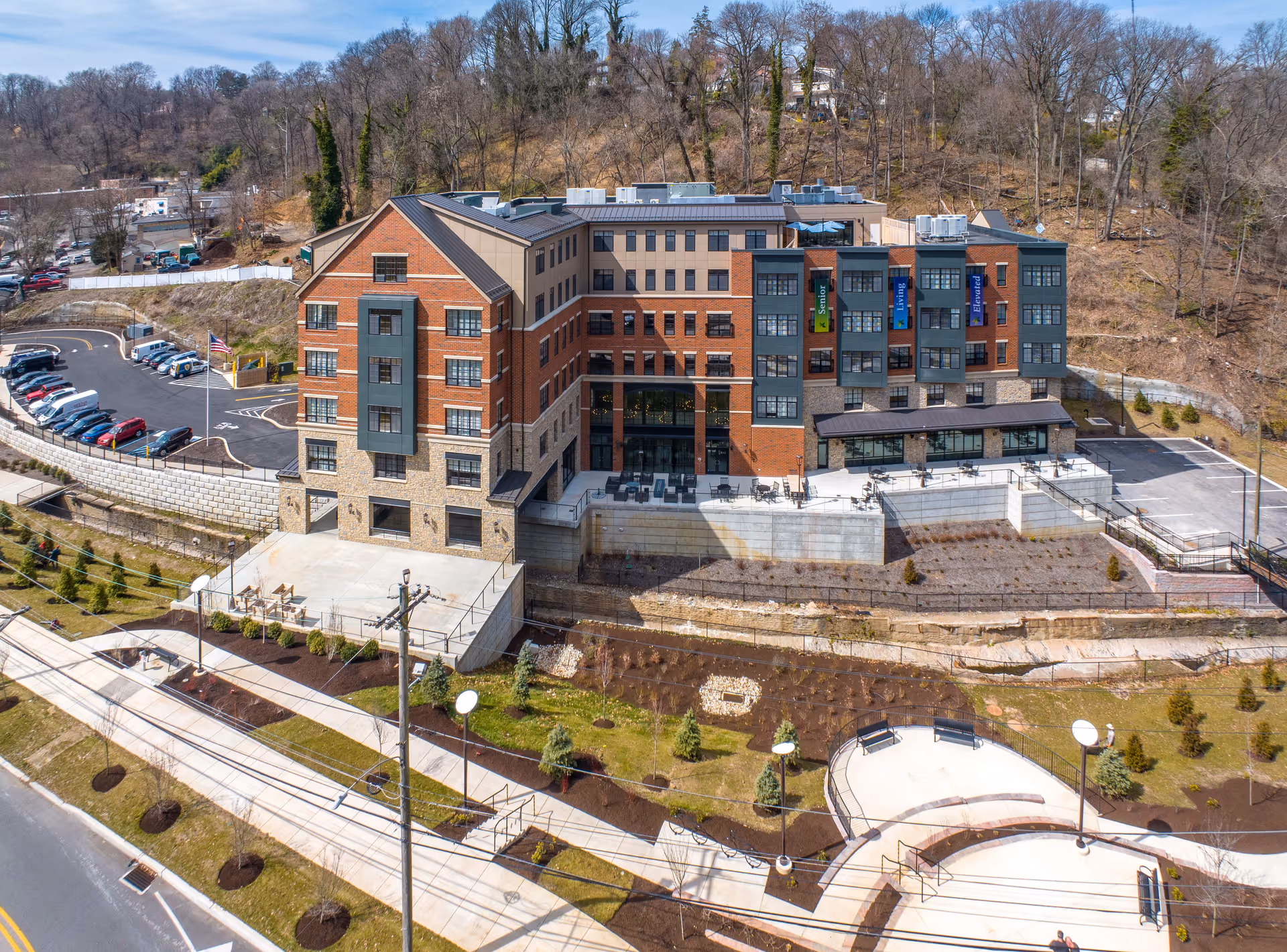 Aerial view of a multi-story senior living facility named The Residence at Bala Cynwyd, featuring a brick and stone exterior with large windows. The building is surrounded by landscaped grounds with small trees, benches, and walking paths. There is a parking lot adjacent to the building and a wooded hill in the background under a clear blue sky.