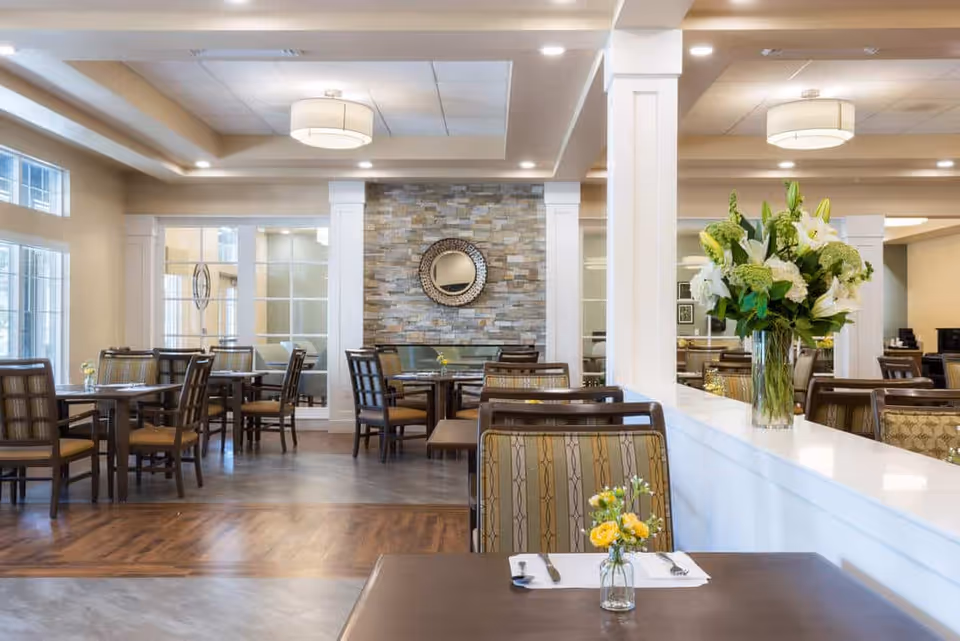A bright and spacious dining room with multiple tables and chairs arranged neatly. The room features large windows allowing natural light to fill the space, a stone accent wall with a round decorative mirror, and ceiling lights. A vase with white and green flowers is placed on a white divider, and a small vase with yellow flowers is on a table in the foreground.
