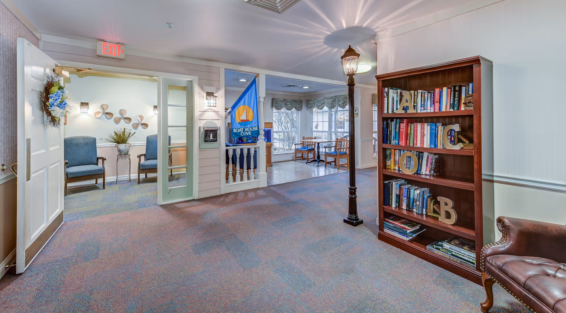 Carpeted senior living facility common area with a bookshelf, seating, a decorative lamp post light, and an open doorway to a sunlit sitting area.