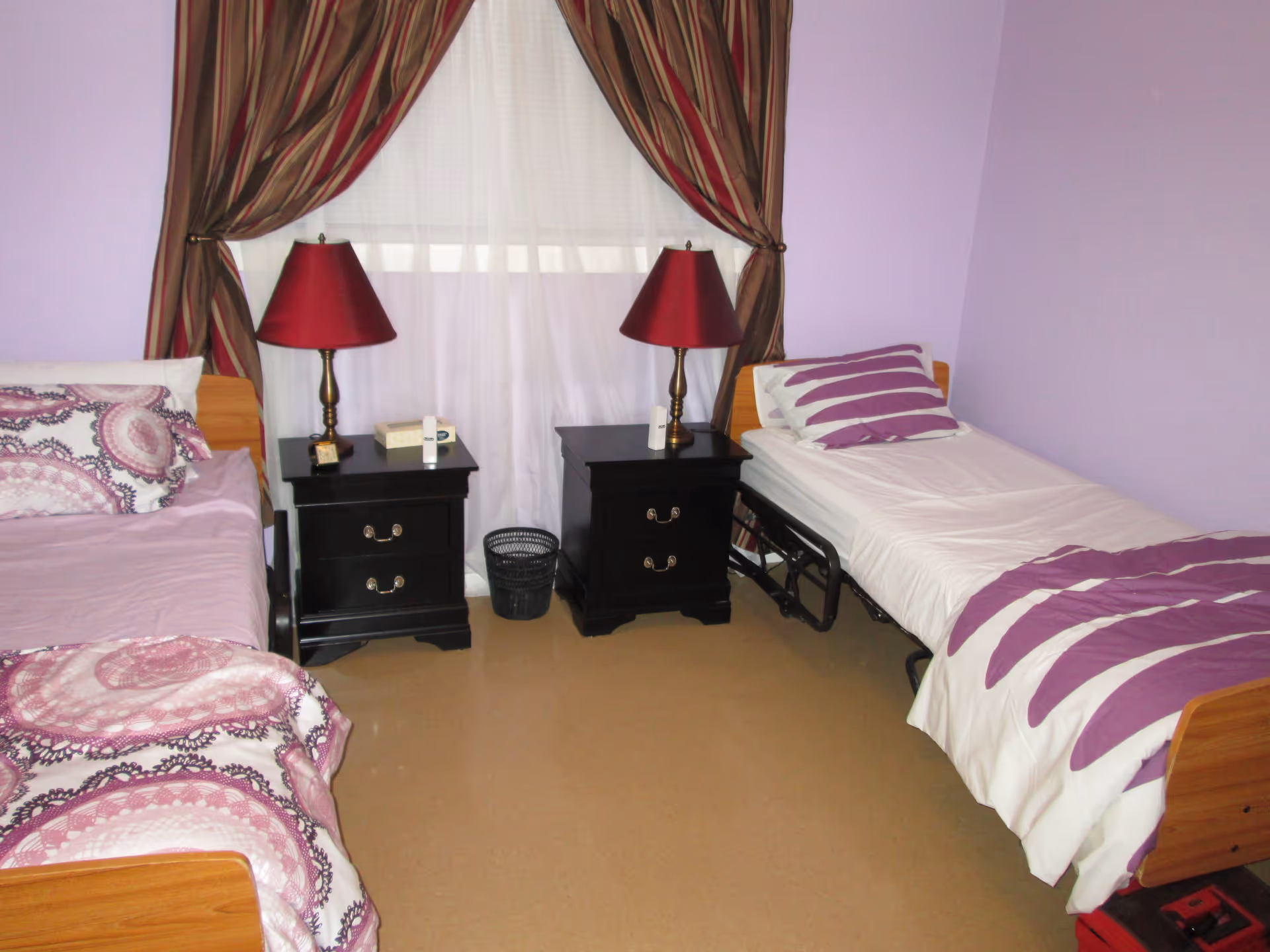A bedroom with two single beds separated by two black nightstands, each with a red lamp. The beds have purple and white bedding, and the walls are painted light purple. A window with sheer white curtains and brown striped drapes is behind the nightstands.