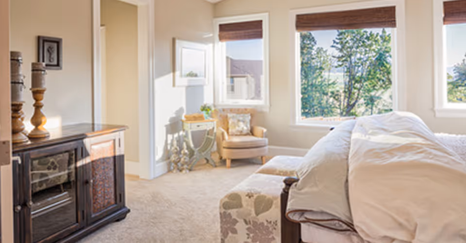 A bright bedroom with a large bed covered in white bedding, a floral-patterned bench at the foot of the bed, a wooden cabinet with decorative items on top, a beige armchair with a pillow, a small side table, and large windows showing green trees outside.