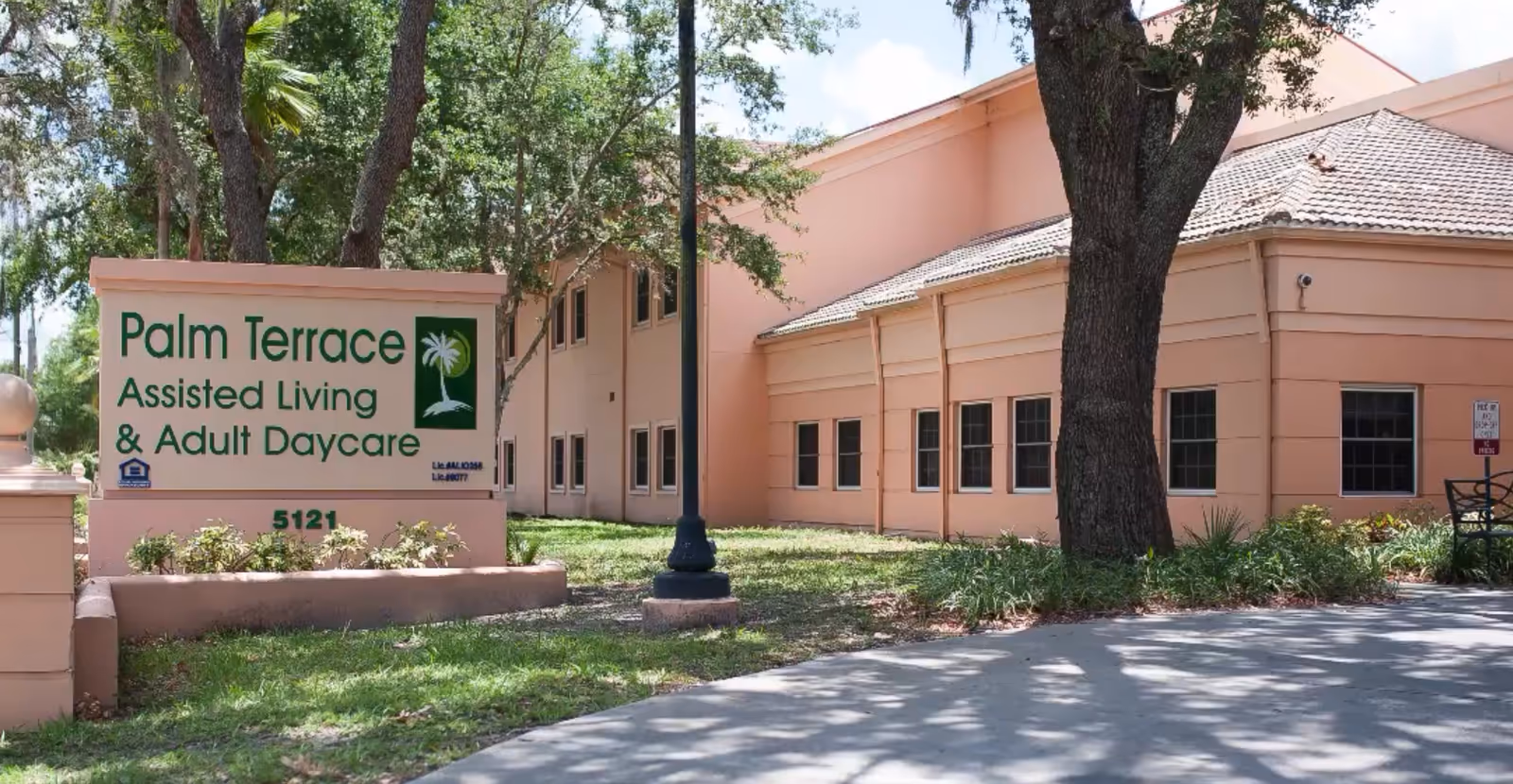 Exterior front of Palm Terrace Assisted Living & Adult Daycare showing the building, large entrance sign, trees, and landscaping.