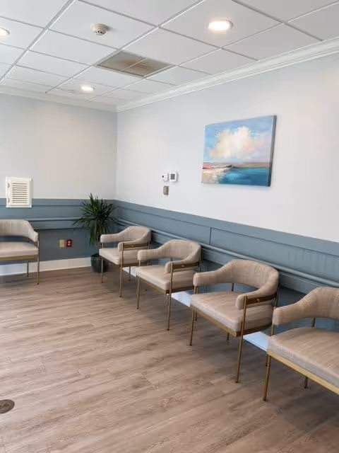 A waiting area with six beige cushioned chairs with gold metal frames arranged along two walls. The room has light wood flooring, light gray walls with blue wainscoting, a potted plant in the corner, and a painting of a cloudy sky over water hanging on the wall. The ceiling has recessed lighting and a smoke detector.