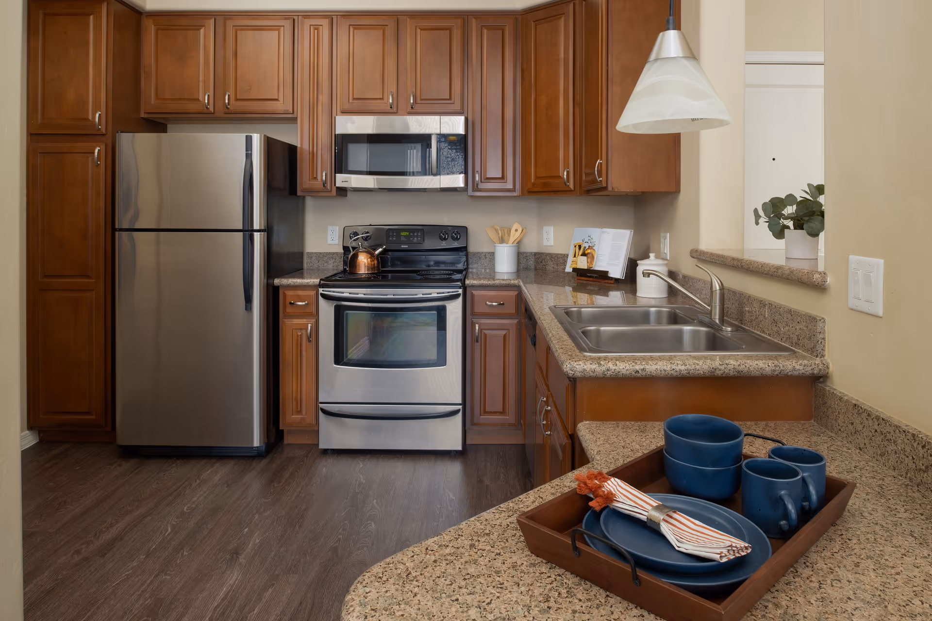 A modern kitchen with wooden cabinets, stainless steel refrigerator, stove, and microwave. The kitchen features granite countertops with a double sink, a hanging light fixture, and a tray with blue dishes and napkins on the counter.