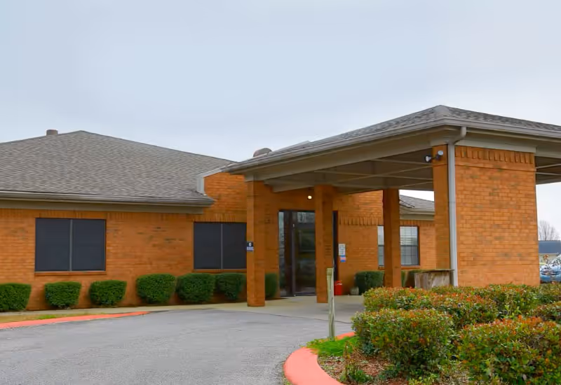 Exterior view of a single-story brick building with a covered entrance supported by brick columns. There are trimmed bushes along the building and a curved driveway leading to the entrance under an overhang.