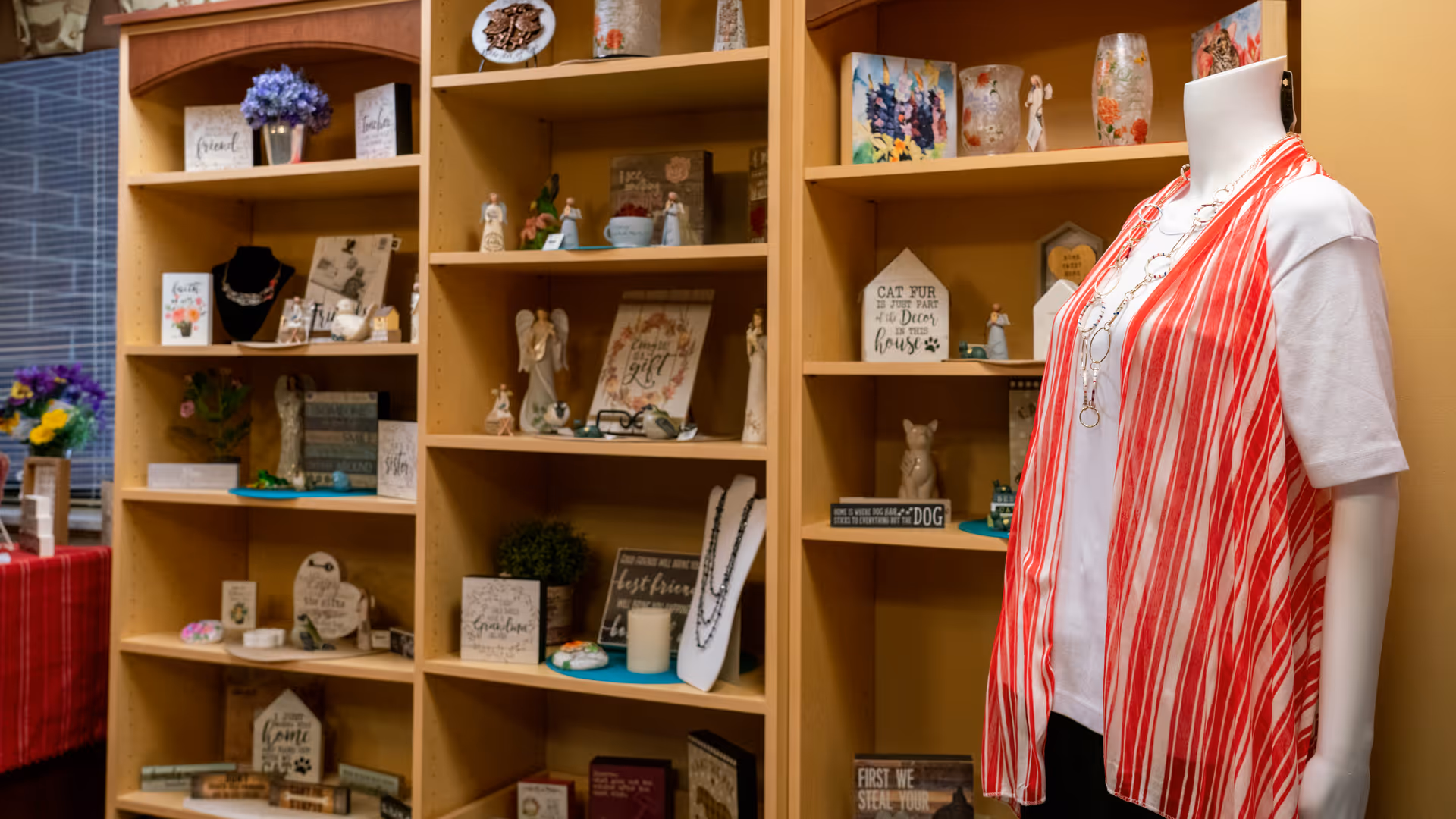 Interior view of a room with wooden shelves filled with decorative items such as figurines, framed quotes, candles, and small plants. To the right, there is a mannequin dressed in a white shirt and a red and white striped vest with a long necklace. A window with blinds and a table with a red checkered cloth and flower arrangements are visible on the left side.