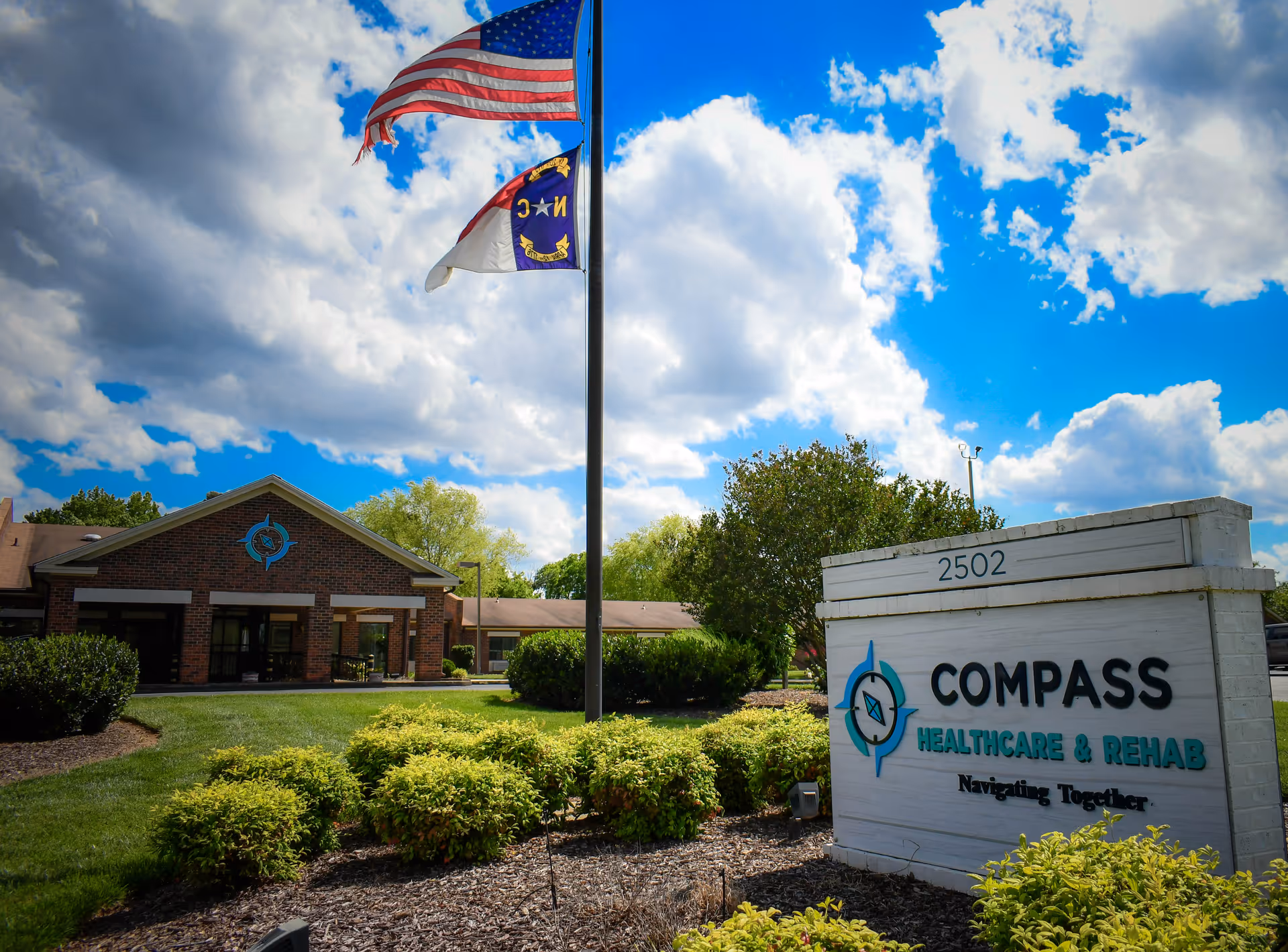 Exterior view of Compass Healthcare and Rehab Hawfields facility with a brick building, a flagpole displaying the American and North Carolina flags, and a white sign with the facility's name and address 2502. The sky is partly cloudy with blue patches.