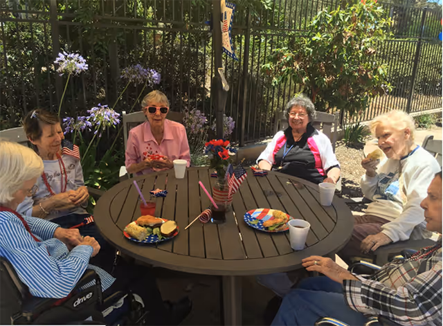 A group of six elderly women sitting around a round outdoor table enjoying a meal together. The table has plates with sandwiches and drinks, small American flags, and a small flower arrangement. They are seated in a garden area with greenery and a metal fence in the background, and the weather appears sunny.