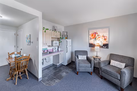 A cozy assisted living apartment interior featuring a small kitchen area with white cabinets, a microwave, and a refrigerator. Adjacent to the kitchen is a dining area with a wooden table and chairs. The living area includes two gray upholstered chairs, a small side table with a lamp, and a floral wall art piece above the chairs. The flooring transitions from tile in the kitchen to carpet in the living and dining areas.