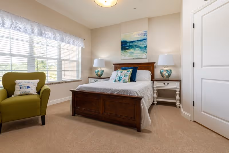 A bright furnished bedroom with a wooden bed, two nightstands and lamps, artwork above the bed, and a green upholstered chair by a large window.