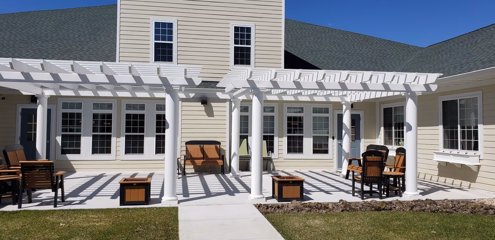 Outdoor patio area at Birchwood Cottages featuring white pergolas with columns, wooden chairs and benches, and a concrete walkway surrounded by grass and landscaping under a clear blue sky.