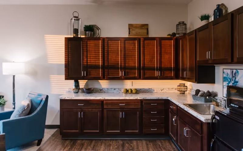 A kitchen area with dark wooden cabinets and a marble countertop. There is a stainless steel sink, a microwave, and decorative items on the counter and above the cabinets. To the left, a blue armchair and a floor lamp are partially visible.