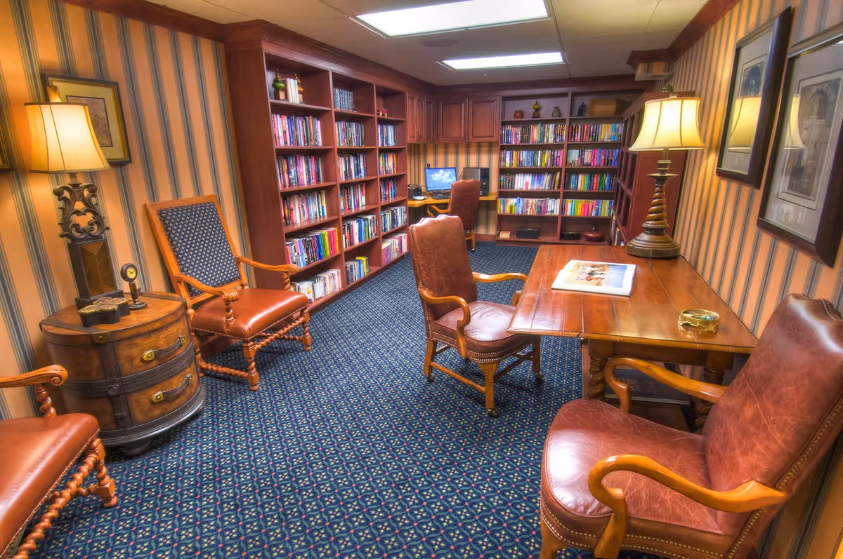 A cozy library room with wooden bookshelves filled with books along the walls, a wooden table with leather chairs around it, a computer desk with a chair in the back corner, patterned carpet, striped wallpaper, and two table lamps providing warm lighting.