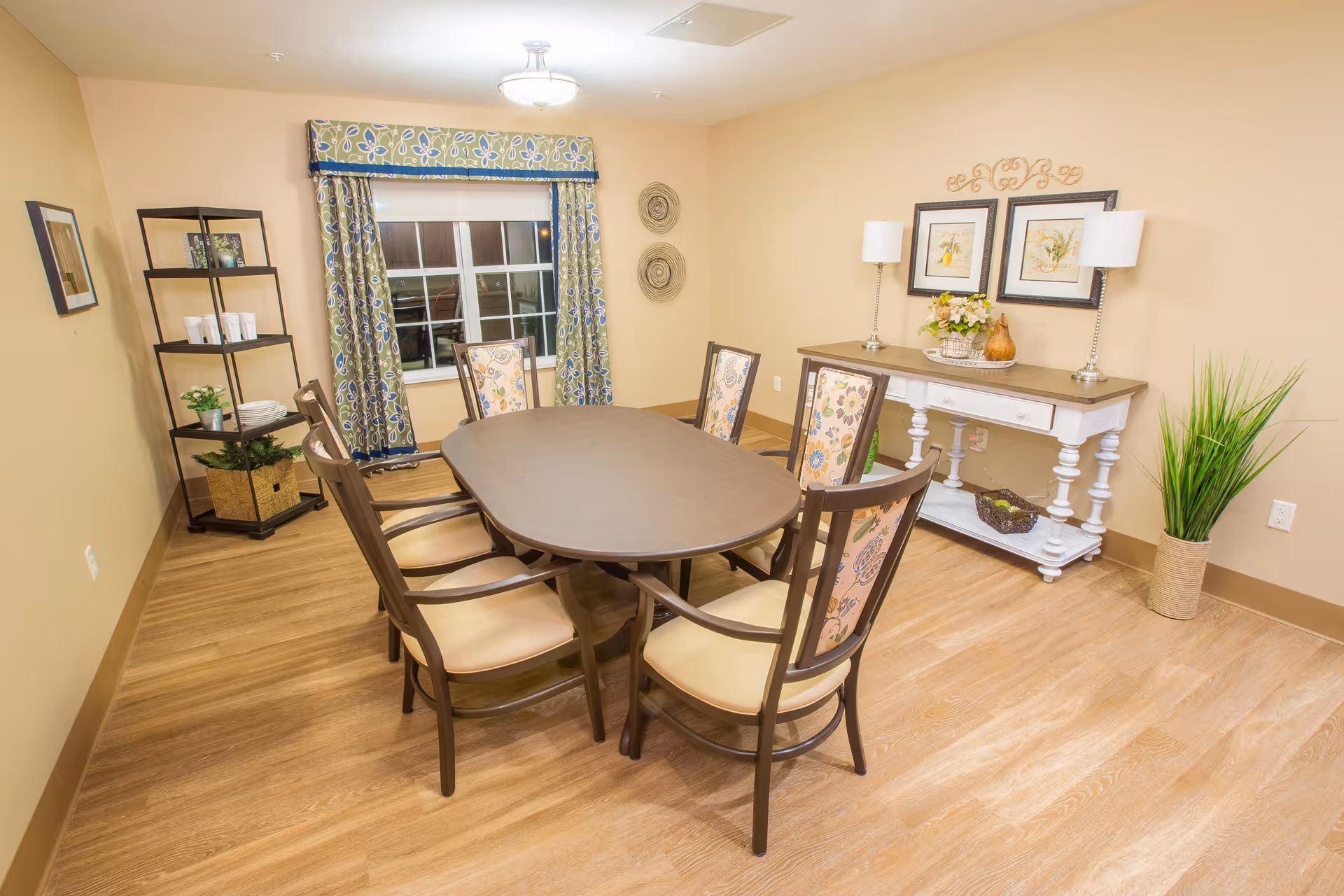 A well-lit dining room with a wooden oval table surrounded by six chairs with floral-patterned backs and beige seats. The room has light-colored walls and wood flooring. A window with floral curtains is on one wall, and a white console table with two lamps, framed artwork, and decorative items is against another wall. A black shelving unit with plants and cups is in the corner, and a tall green plant in a woven basket is near the console table.