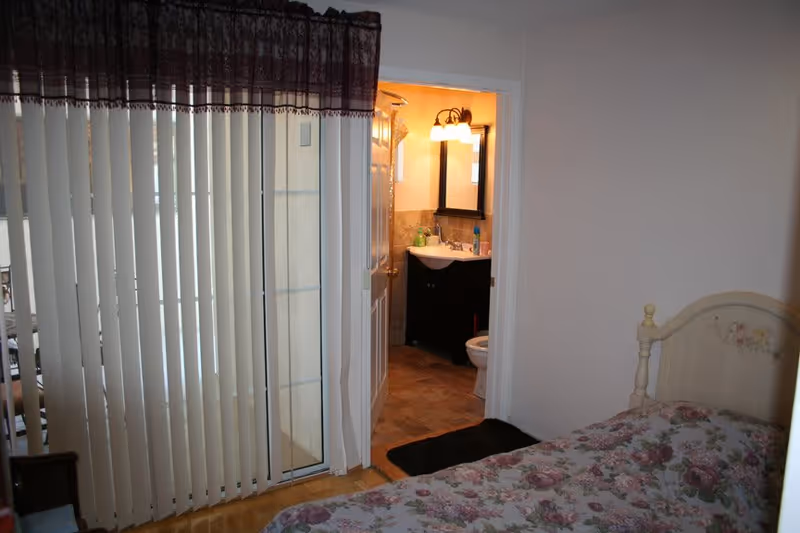 View of a bedroom with a floral bedspread on a bed, vertical blinds covering a sliding glass door, and an open door leading to a bathroom with a sink, mirror, and toilet visible.
