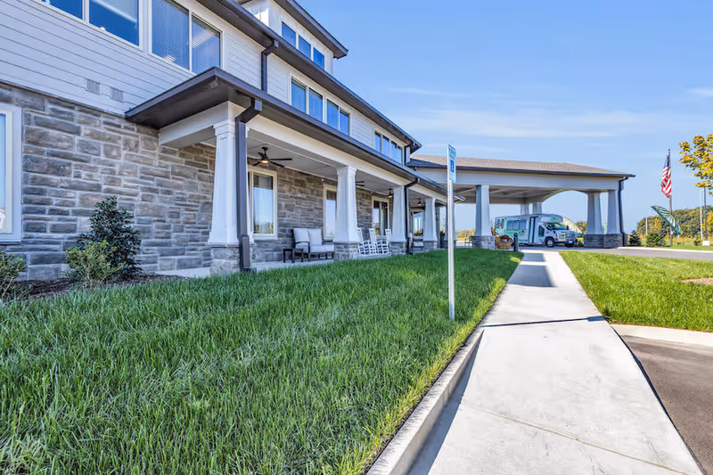 Exterior view of a senior living facility with a covered entrance supported by white columns, a sidewalk leading to the entrance, green grass, and a vehicle parked under the covered area. The sky is clear and blue.