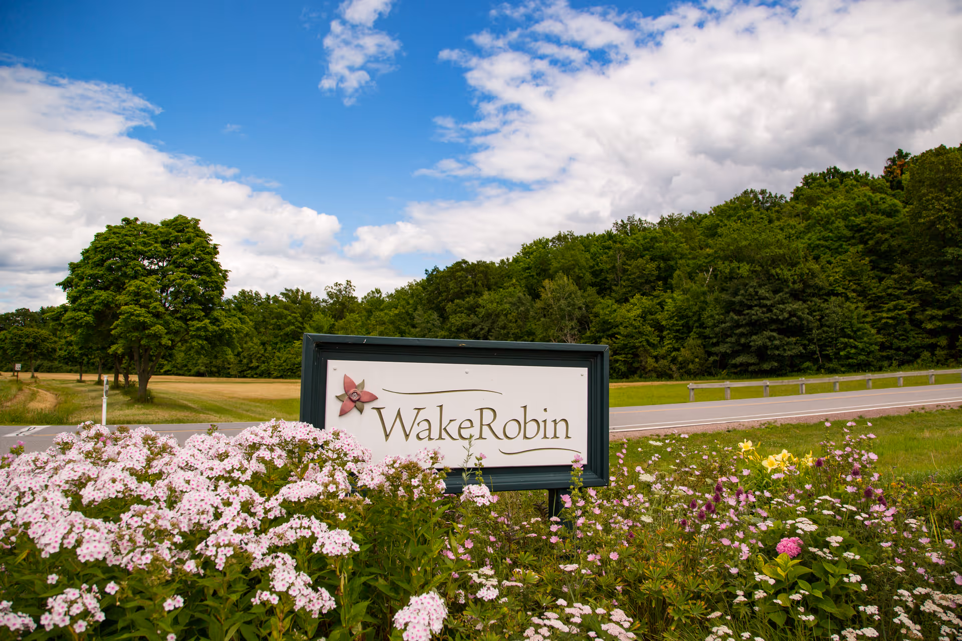 A sign reading 'Wake Robin' surrounded by blooming flowers with a road and green trees in the background under a partly cloudy blue sky.