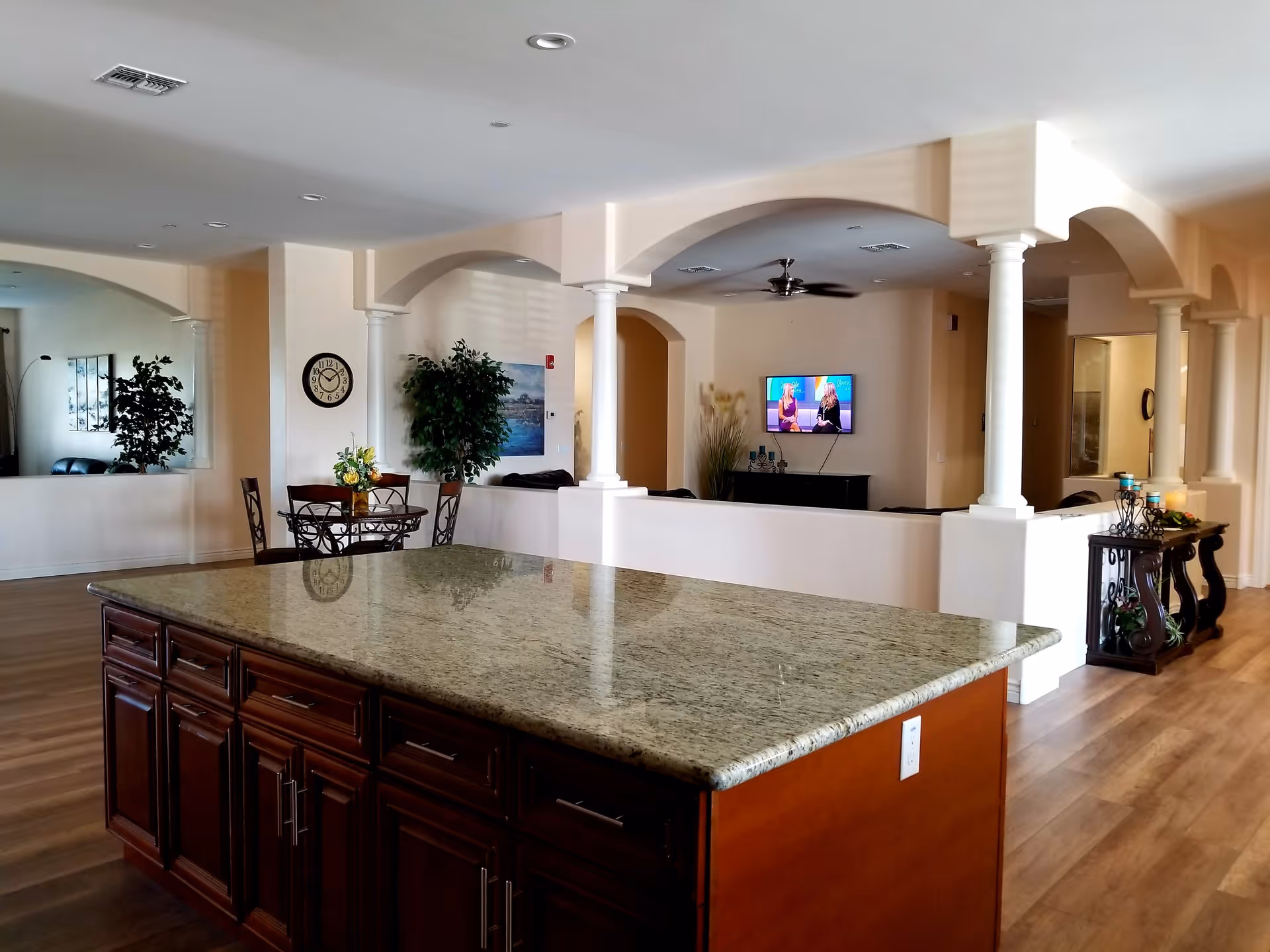 Interior view of a senior living facility common area featuring a large granite kitchen island with wooden cabinets, a small dining table with chairs, decorative plants, a wall clock, and a television mounted on the wall in a lounge area with arched openings and columns.