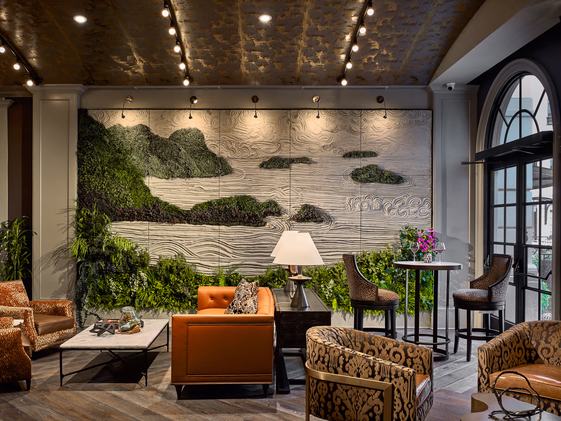 A stylish living room area featuring a large textured wall art with green foliage, a brown leather sofa, patterned armchairs, a marble coffee table, a side table with a lamp, and a small round table with two chairs near a large arched window.