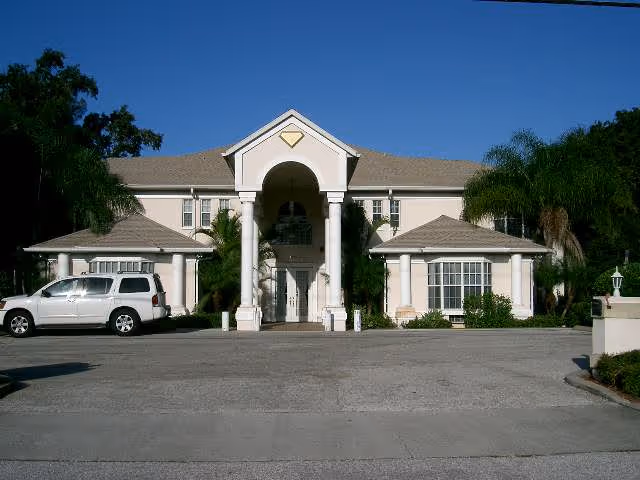 Front exterior view of a large two-story building with a central entrance featuring tall white columns and a triangular pediment. There are two smaller wings on either side of the entrance, palm trees and other greenery around the building, and a silver SUV parked on the left side of the driveway under a clear blue sky.
