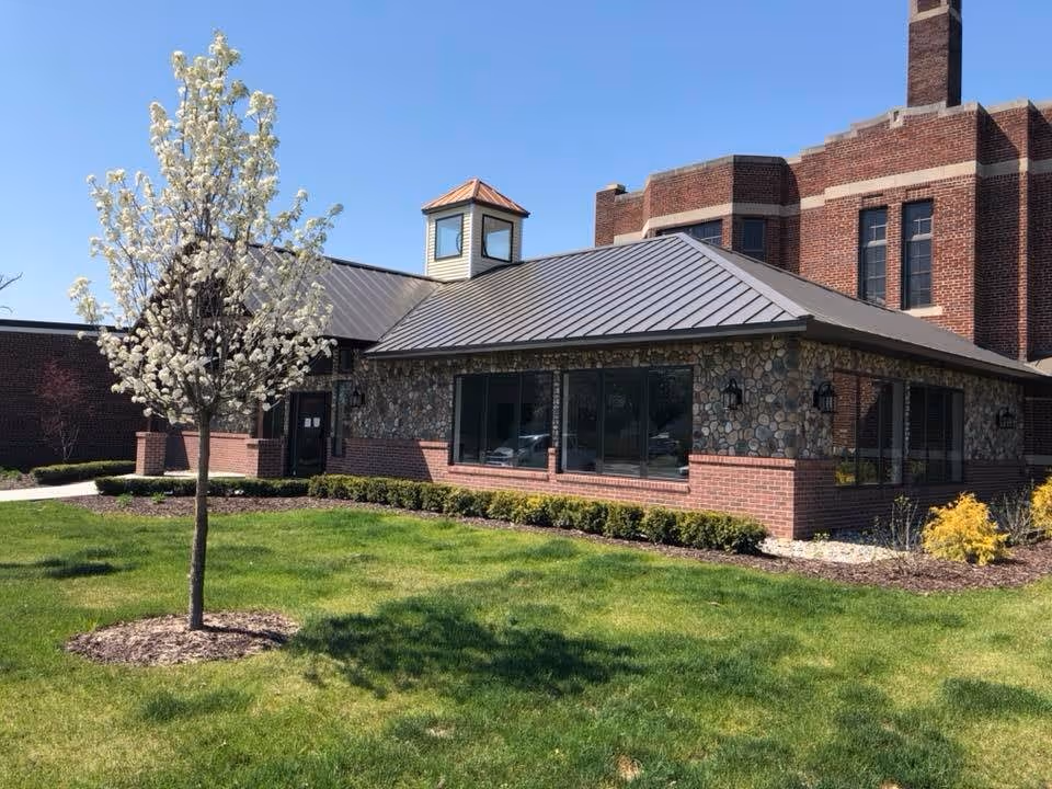 Front exterior of a brick-and-stone senior living building with a metal roof, large windows, and a flowering tree on a grassy lawn.