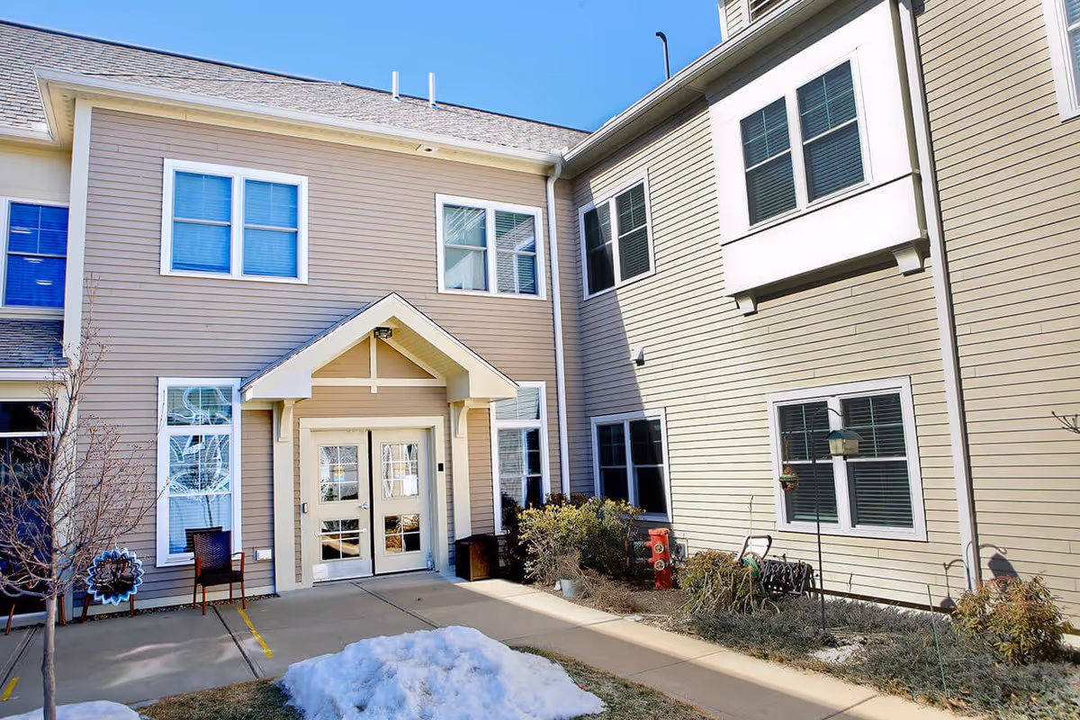 Front entrance of a beige two-story senior living building with double doors, multiple windows, and a small courtyard with patches of snow.