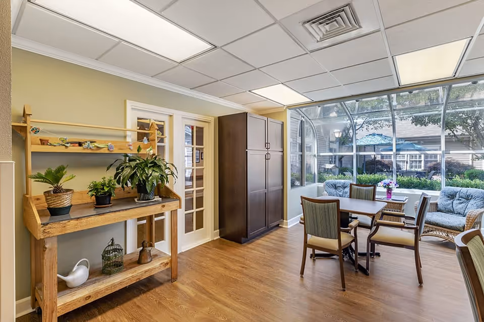 A bright room with wooden flooring featuring a wooden plant stand with potted plants on the left, a dark wooden cabinet in the center, and a seating area with a table, chairs, and cushioned wicker sofas near large windows overlooking an outdoor garden area.