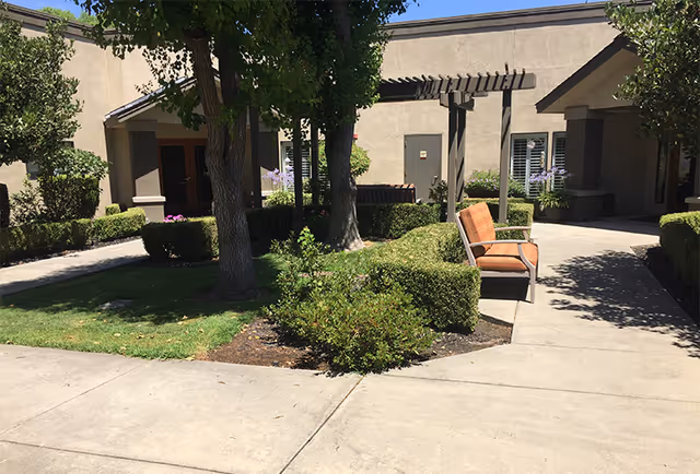 Outdoor courtyard area at Summerfield of Stockton featuring a concrete walkway, trimmed bushes, trees providing shade, and an orange cushioned bench under a pergola attached to a beige building.