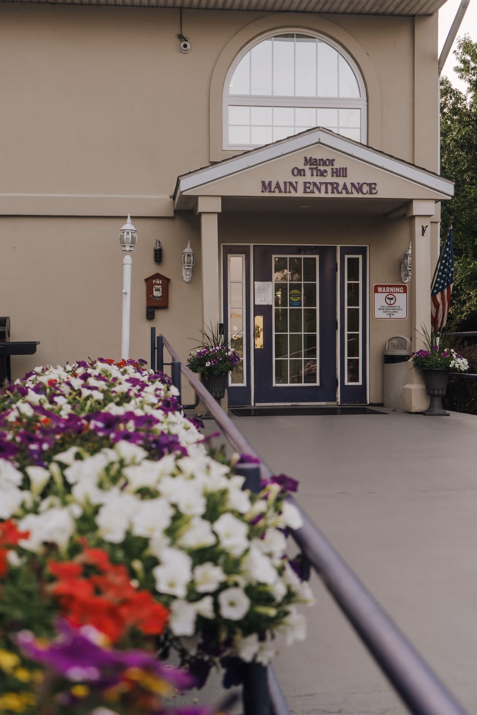 Main entrance of Manor On The Hill building with a ramp leading up to double doors. There are colorful flowers in planters along the ramp railing and two large flower pots on either side of the entrance. An American flag is visible to the right of the entrance.