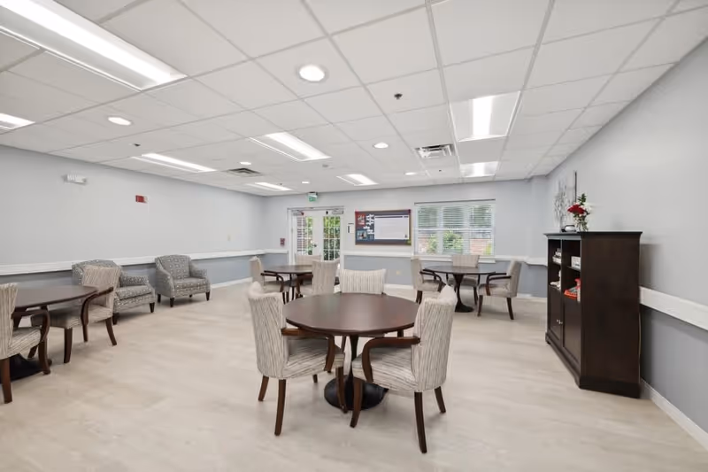 A bright, spacious common area in a senior living community featuring several round wooden tables with upholstered chairs arranged around them. There are additional armchairs along the walls, a bulletin board near a door with glass panels, and a dark wooden cabinet with decorative items and flowers. The room has light-colored flooring, pale gray walls, and a white drop ceiling with recessed lighting.