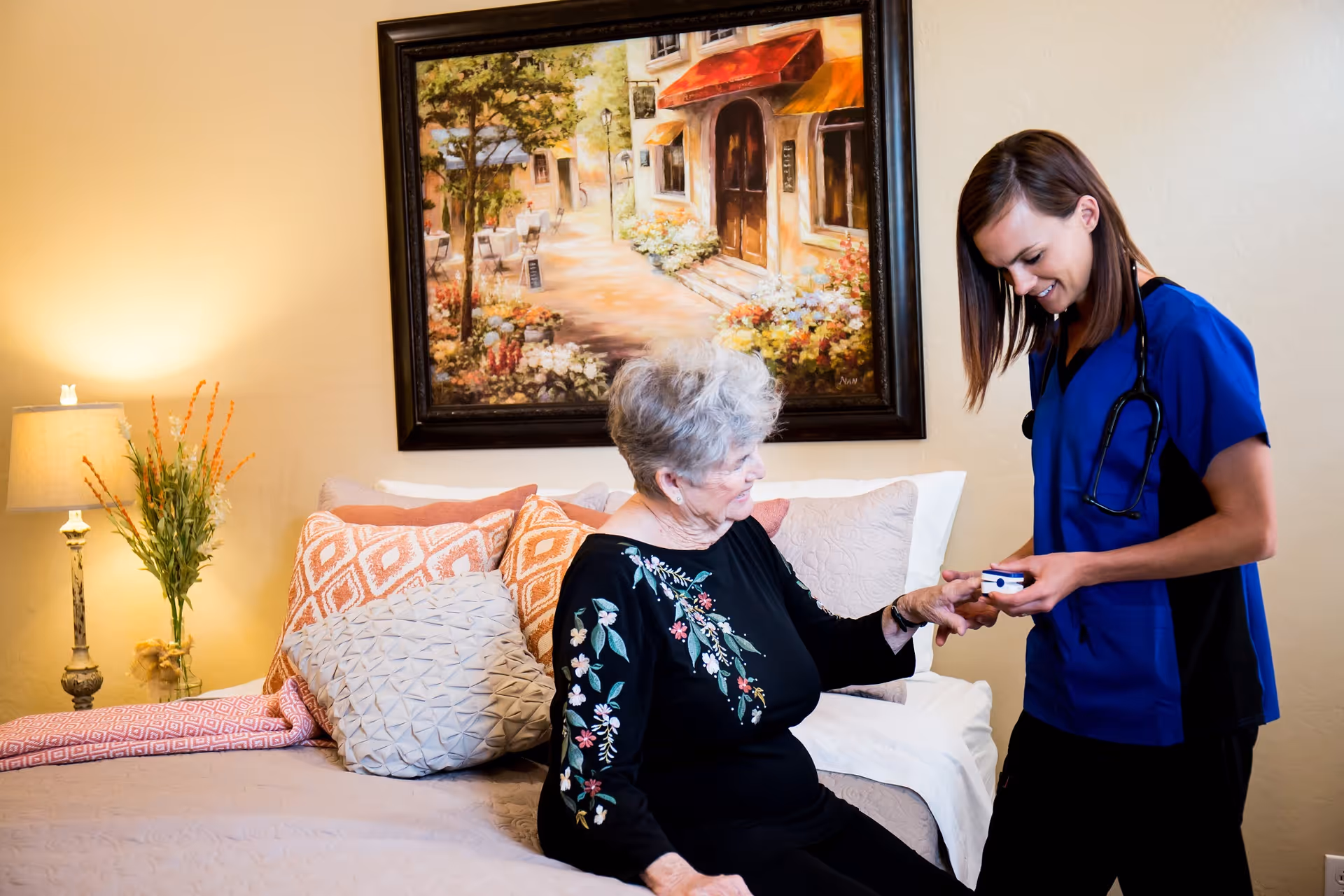 An elderly woman sitting on a bed in a cozy bedroom with decorative pillows and a bedside lamp, while a healthcare professional in blue scrubs with a stethoscope checks her finger with a medical device. A framed painting of a street scene hangs on the wall behind them.