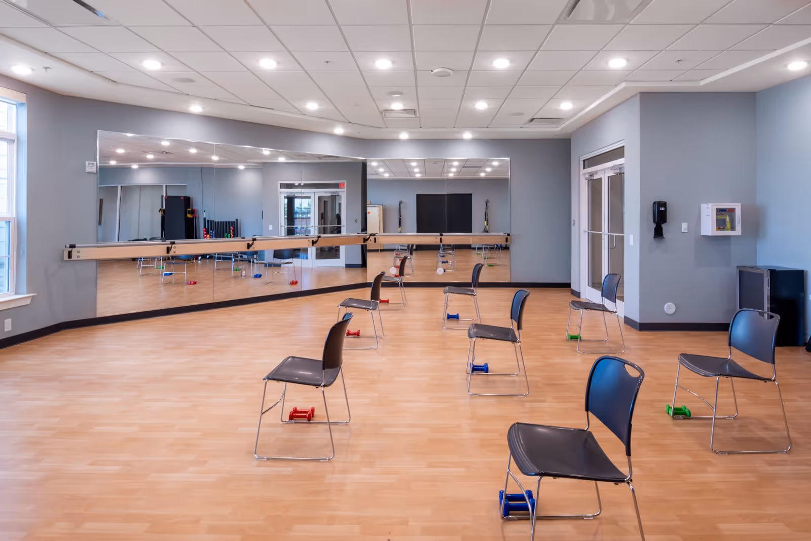 A spacious exercise room with wooden flooring and light gray walls. The room features a large wall mirror with a ballet barre, several black chairs spaced apart, and small colorful dumbbells placed near each chair. The ceiling has recessed lighting, and there are windows on the left side letting in natural light. A door and some exercise equipment are visible in the background.