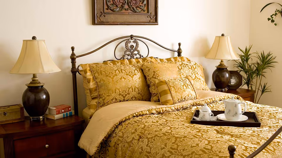 A bedroom with a decorative metal headboard and a bed covered in gold-patterned bedding and pillows. On either side of the bed are wooden nightstands with dark brown lamps and various items including books and a clock. A tray with a teapot and cup is placed on the bed. A green plant is visible in the corner of the room.