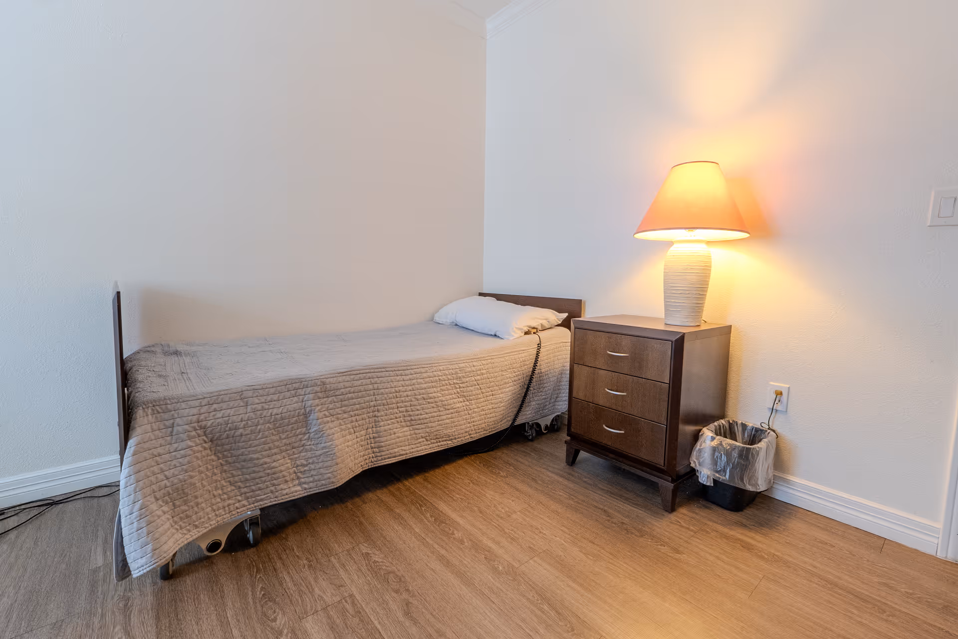 Small bedroom with a single bed, a three-drawer nightstand topped by a lit lamp, and a trash can on wood flooring.