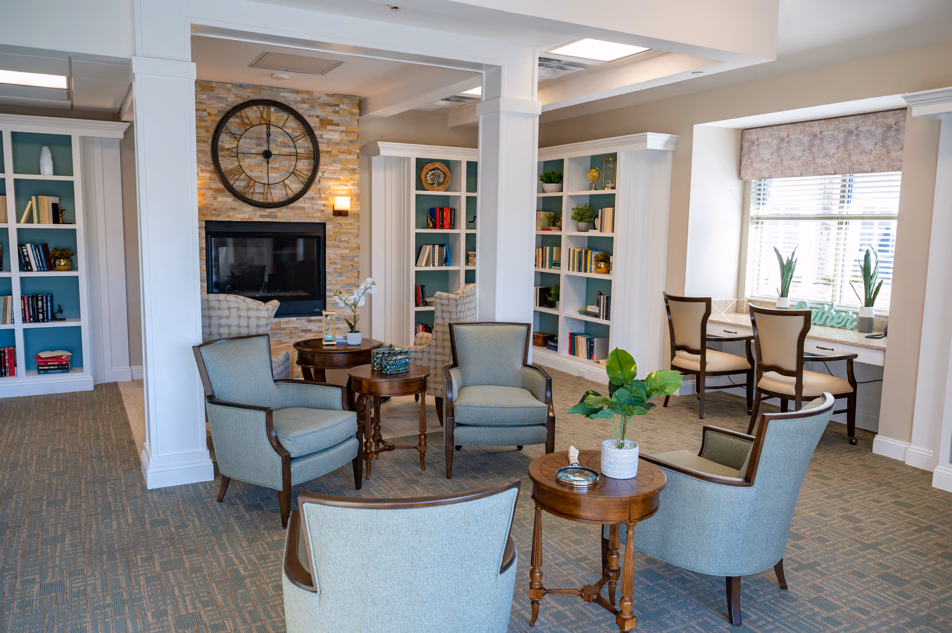 A cozy living room area in a senior living facility with four upholstered armchairs arranged around two small wooden tables. The room features built-in bookshelves filled with books and decorative items, a stone fireplace with a large clock above it, and a window nook with two chairs and a desk. The space is well-lit with natural light coming through the window.