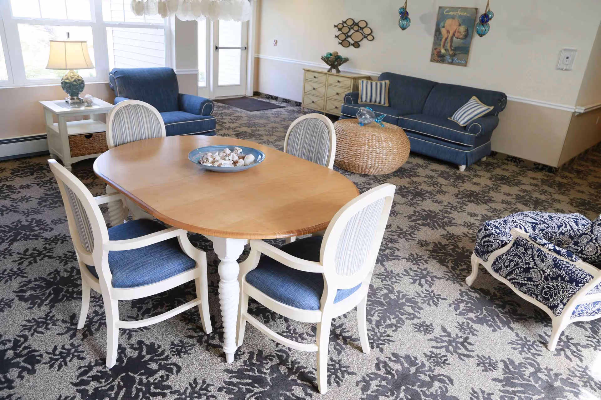 A cozy living room area with a wooden oval table surrounded by four white chairs with blue cushions. Behind the table, there is a blue armchair next to a white side table with a lamp. In the background, a blue sofa with striped pillows and a round wicker coffee table are visible. The carpet has a floral pattern, and the walls are decorated with a wall art piece and hanging glass ornaments. Large windows allow natural light to fill the room.