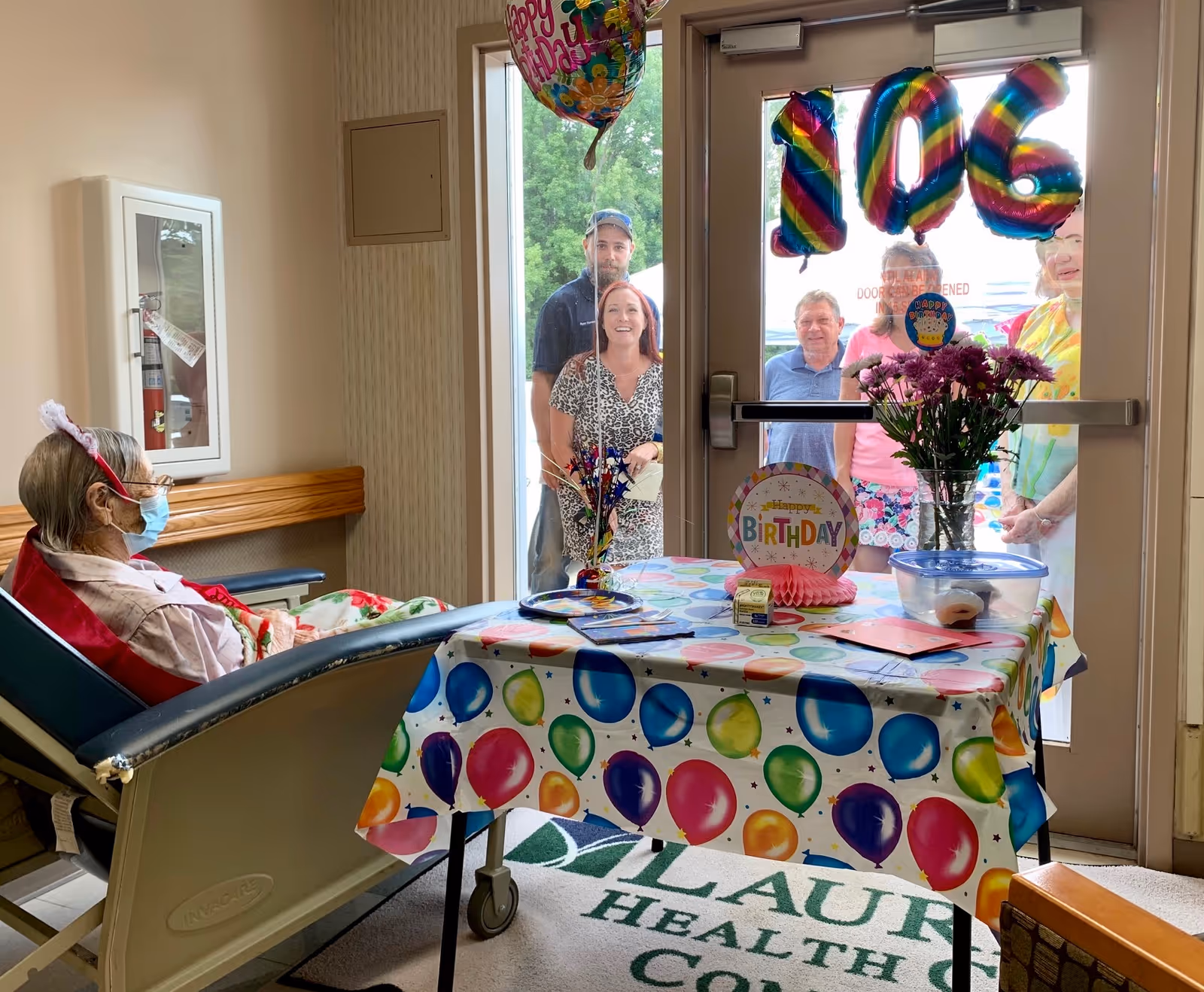 An elderly woman wearing a face mask and birthday headband sits in a wheelchair inside a room decorated for a birthday celebration. A table covered with a colorful balloon-patterned tablecloth holds birthday decorations, flowers, and cards. Outside the glass door, several people stand smiling, looking in. Large rainbow-colored balloons on the door display the number 106.