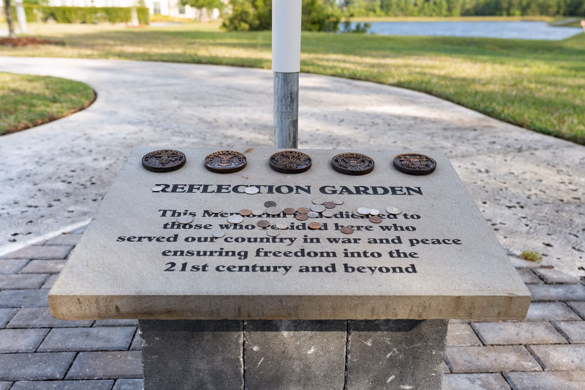 A stone memorial plaque labeled "REFLECTION GARDEN" with military medallions and coins on top, set on a pedestal beside a curved walkway and pond.