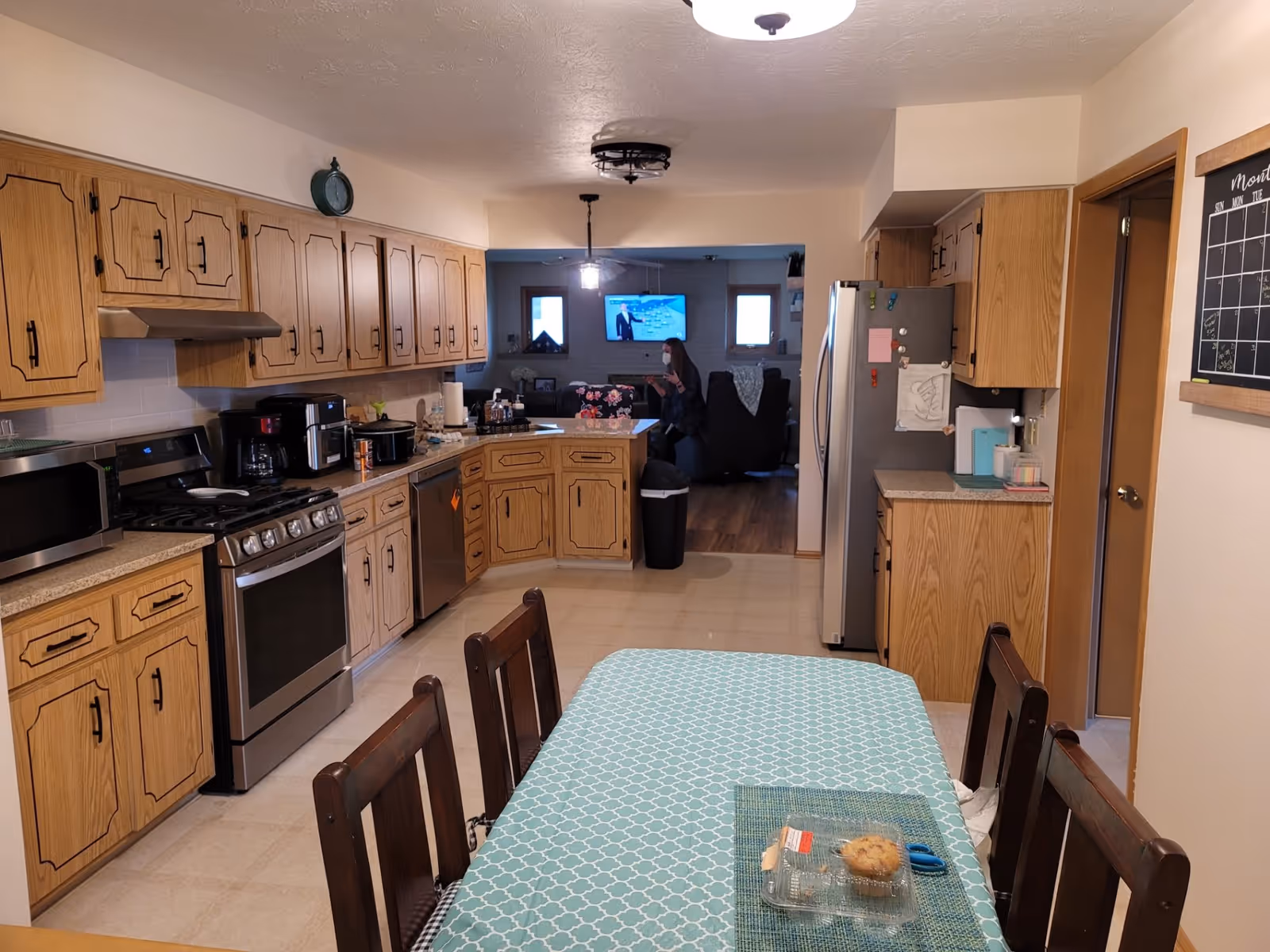 A kitchen with wooden cabinets, stainless steel appliances including a stove, microwave, dishwasher, and refrigerator. A dining table with a blue patterned tablecloth and four wooden chairs is in the foreground. In the background, a living room area is visible with a person wearing a mask sitting on a chair and a TV mounted on the wall.