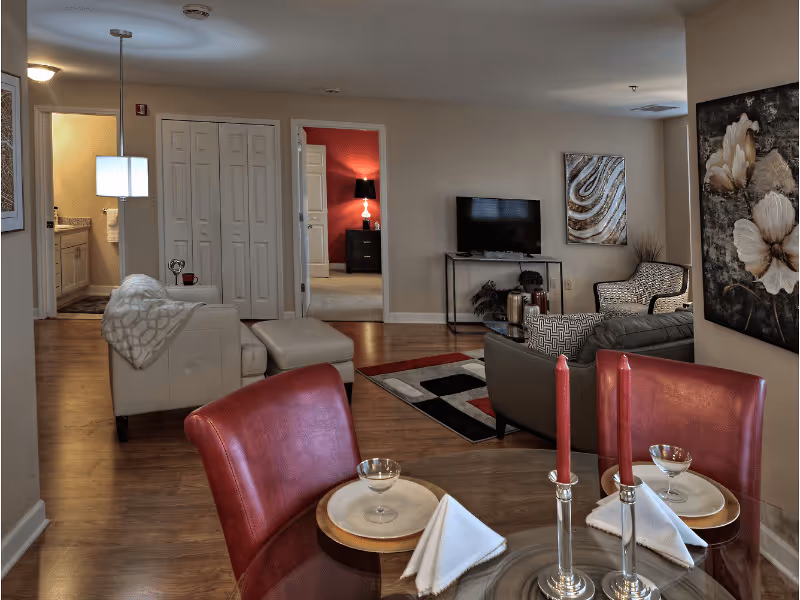 A cozy living and dining area in a senior living facility. The foreground shows a round glass dining table set for two with red leather chairs, white napkins, plates, and two red candles in silver holders. The living room area features a gray sofa, a patterned armchair, a TV on a stand, and decorative wall art. In the background, there is a hallway leading to a bedroom with a red accent wall and a lit lamp on a nightstand. The flooring is wooden throughout.